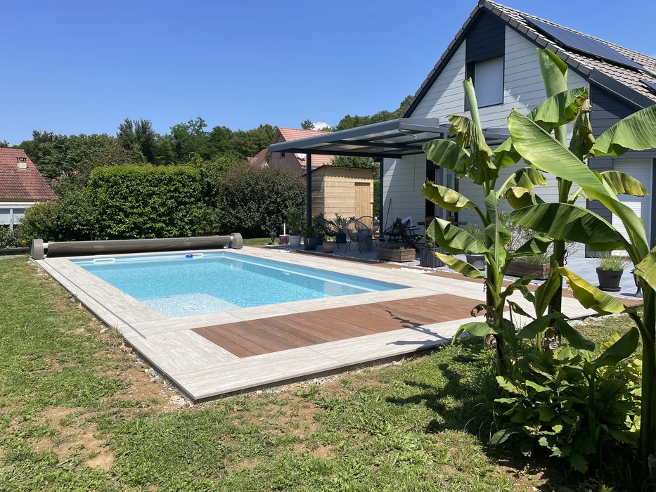 Piscine rectangulaire enterrée avec liner bleu, entourée d'une margelle en carrelage imitation pierre et d'un lé de terrasse en bois composite